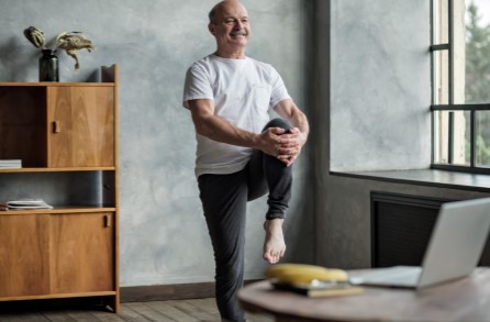 A bald aged man excercising at home in front of a laptop & with one knee raised & held with both hands